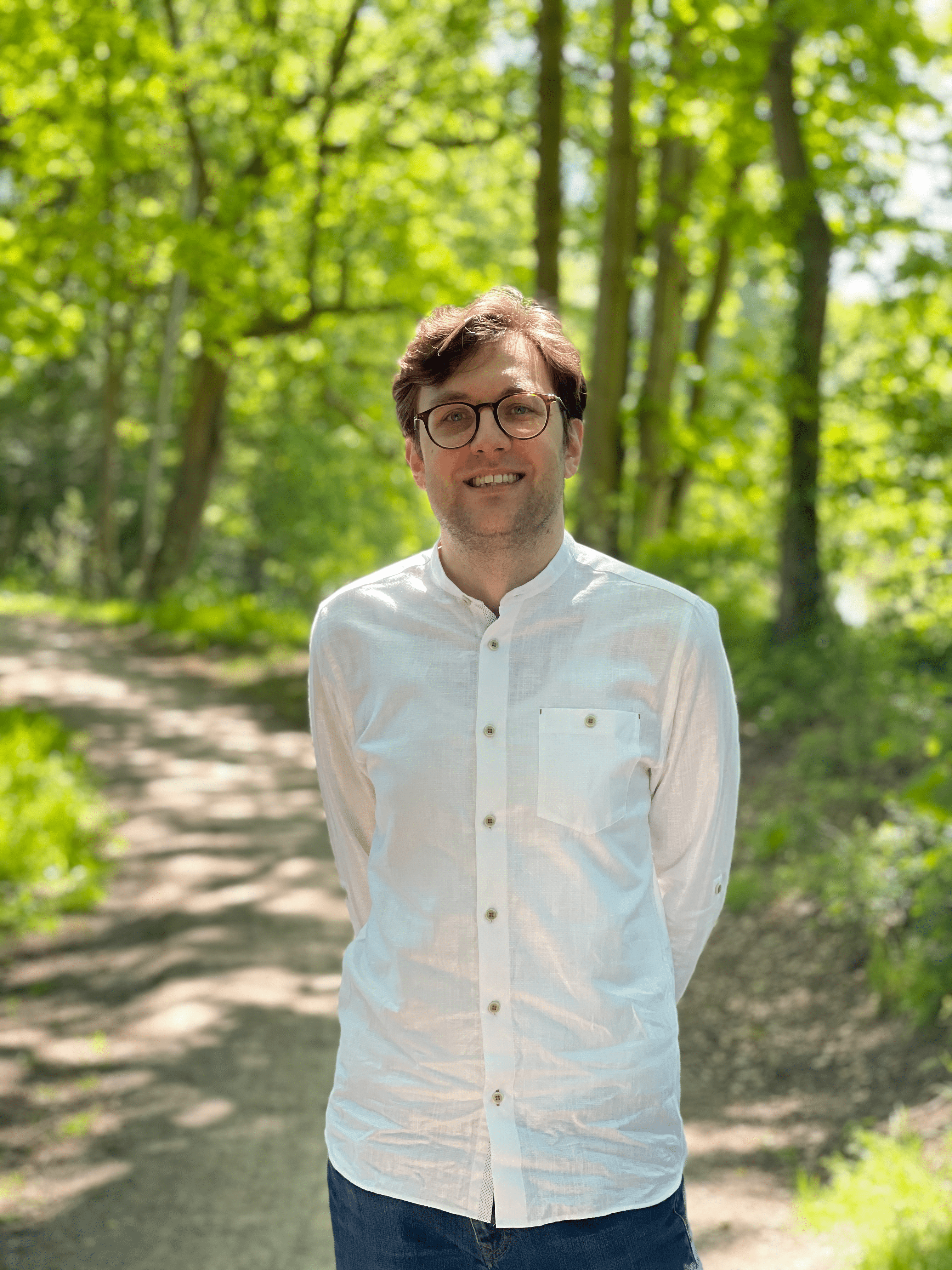 A photo of me, Sander, from the waist up. I'm wearing a white button-down shirt that is untucked, with round, brown glasses. The background consists of a path in a forest-like environment.